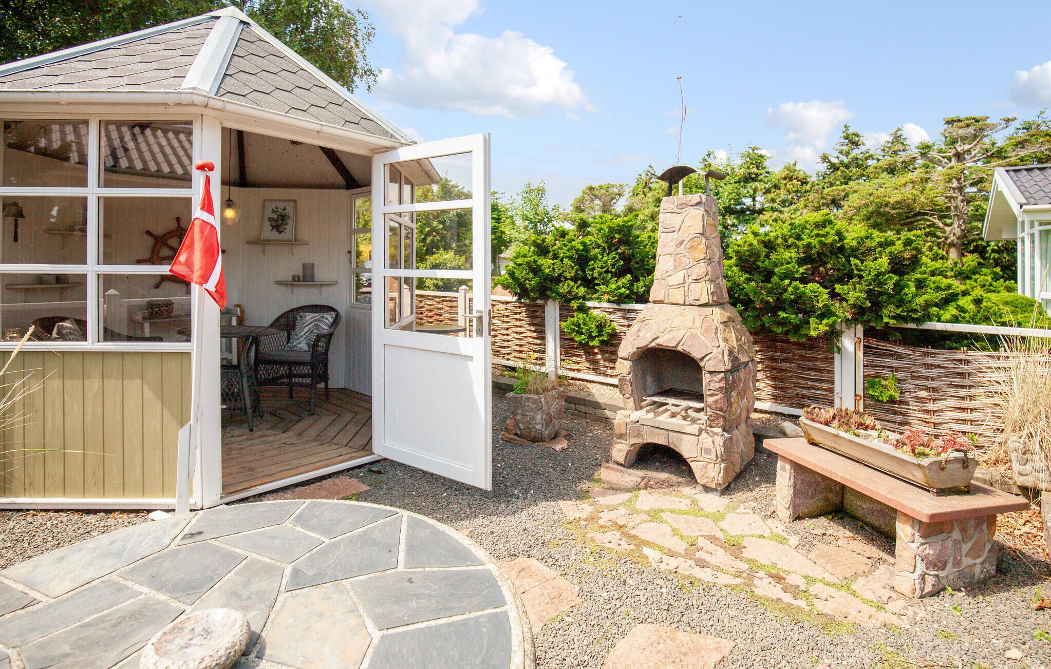 A cozy outdoor seating area with a stone-built oven, a wooden deck, and a view of lush greenery.
