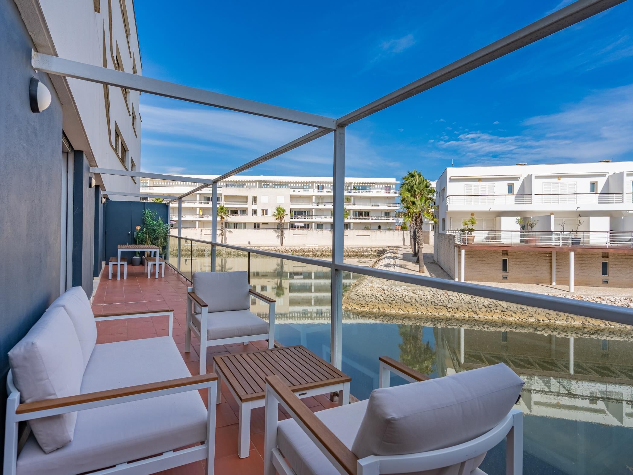 Un balcon doté de mobilier blanc, d’une barrière en verre et offrant une vue sur des immeubles et des palmiers.