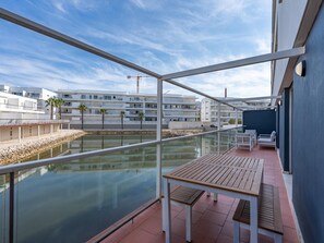 Ein Balkon mit Blick auf ein ruhiges Gewässer, einem Holztisch und einem Glasgeländer.