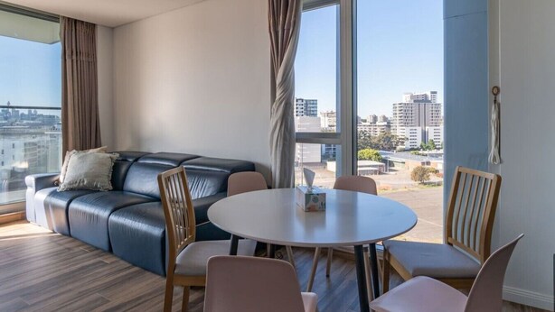 A modern living room with a leather sofa, a round dining table, and chairs. Large windows offer a city view. - City and Park View Close to Station 1 Car Space (Beaconsfield)
