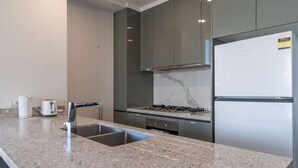 A modern kitchen with a granite countertop, stainless steel sink, gas stove, and a white refrigerator. - City and Park View Close to Station 1 Car Space (Beaconsfield)