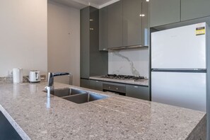 A modern kitchen with a granite countertop, stainless steel sink, gas stove, and a white refrigerator. - City and Park View Close to Station 1 Car Space (Beaconsfield)