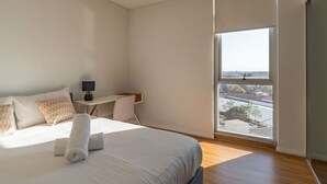 A modern bedroom with a large bed, a desk, and a view of the city through a large window. - City and Park View Close to Station 1 Car Space (Beaconsfield)