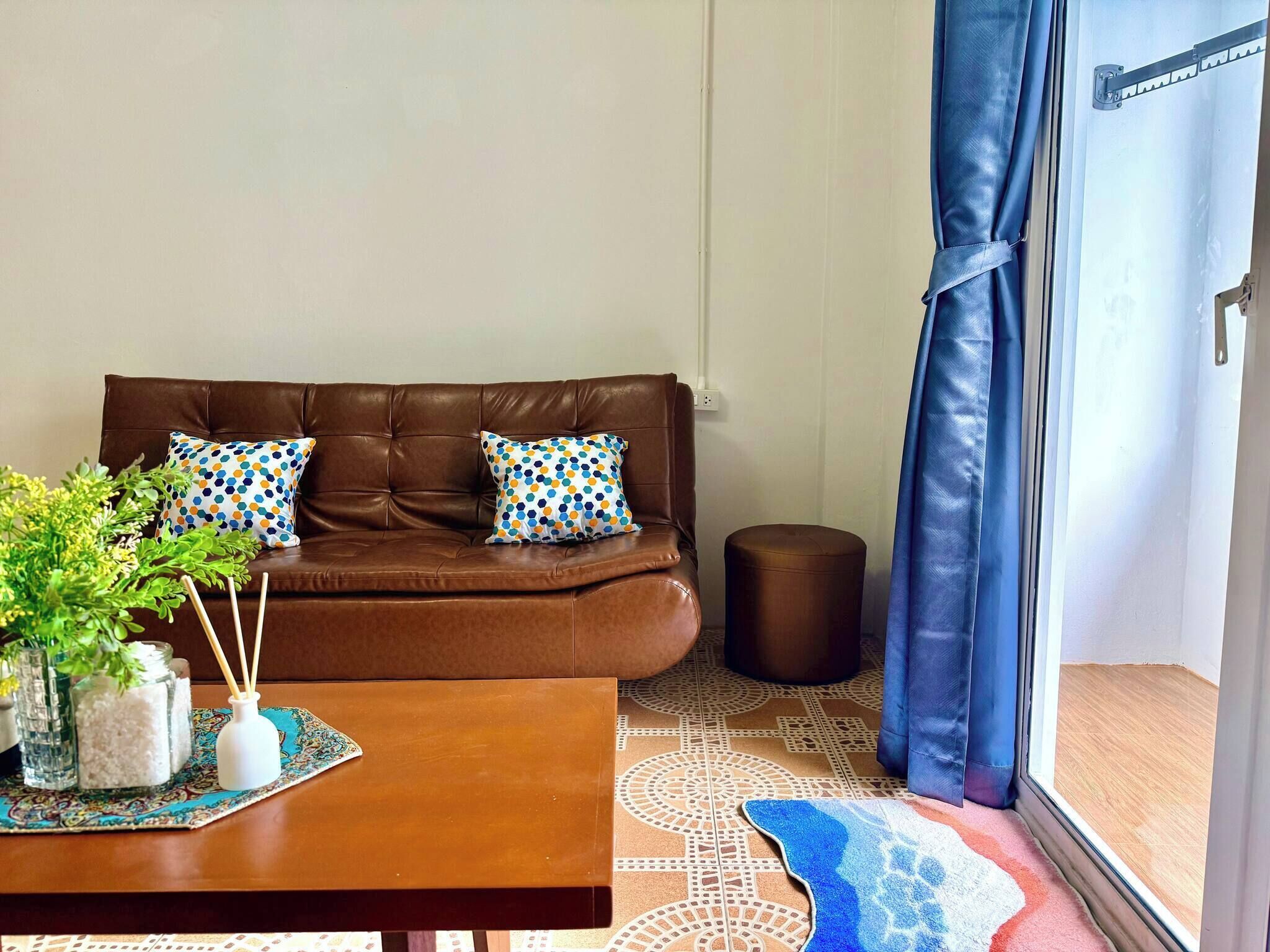 A brown leather sofa with two patterned pillows, a wooden coffee table with a decorative mat, a small round ottoman, and a patterned rug.