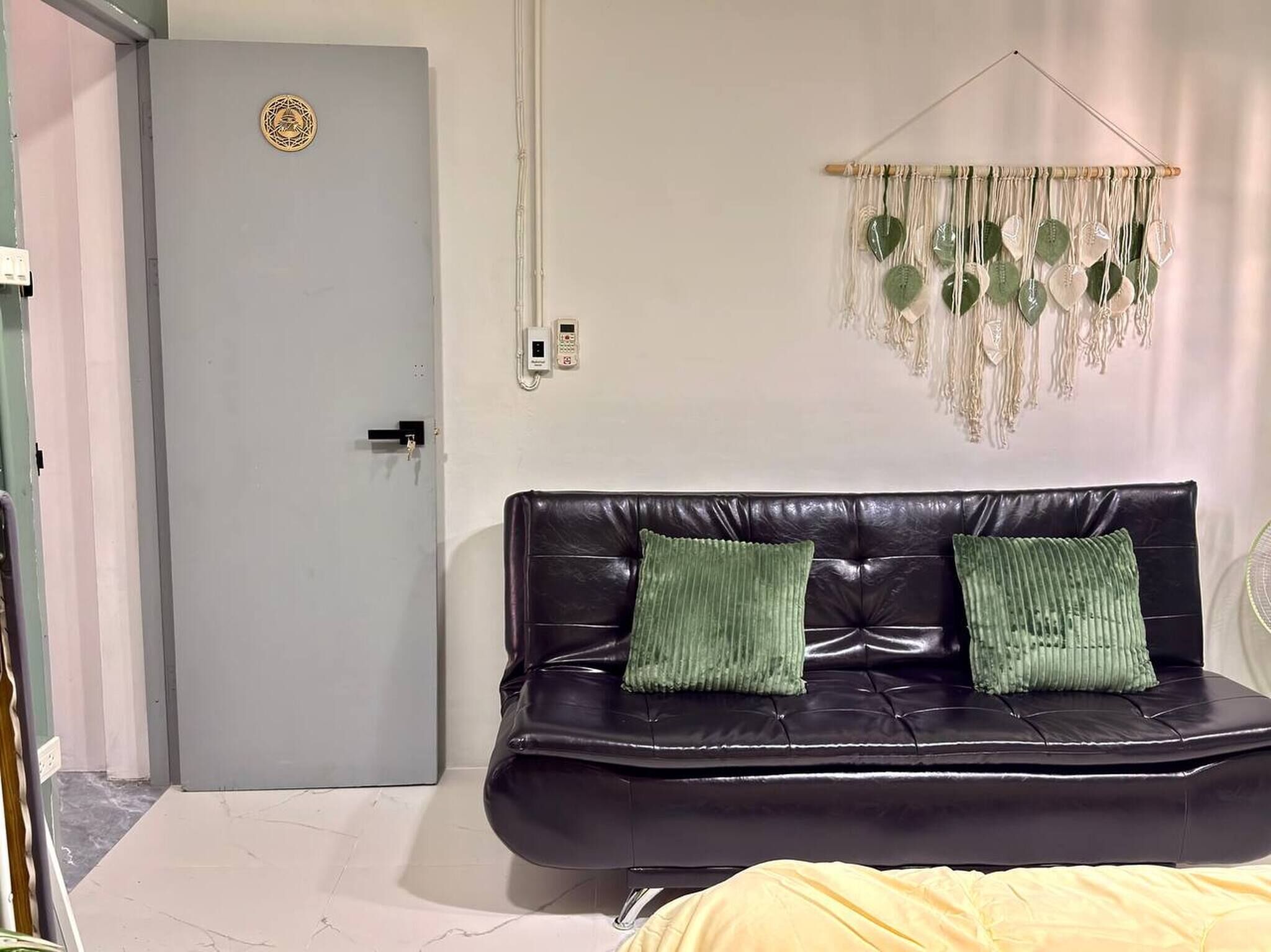 A room with a grey door, a black leather sofa with two green cushions, and a macrame wall hanging.