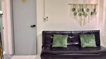 A room with a grey door, a black leather sofa with two green cushions, and a macrame wall hanging.