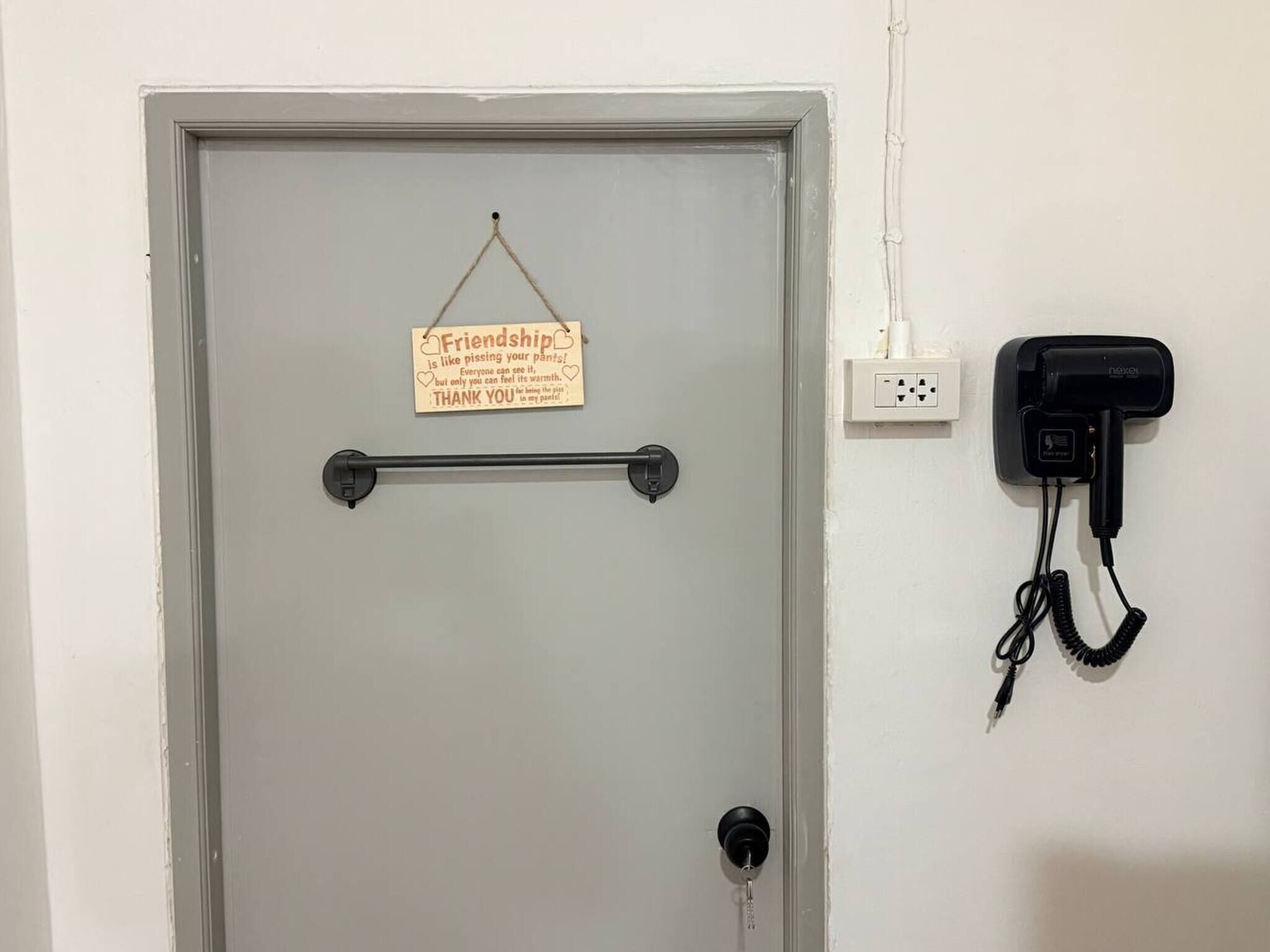 A grey door with a sign that reads 'Friendship is like pissing your pants! Everyone can see it, but only you can feel its warmth. THANK YOU!' A black telephone is mounted on the wall to the right.