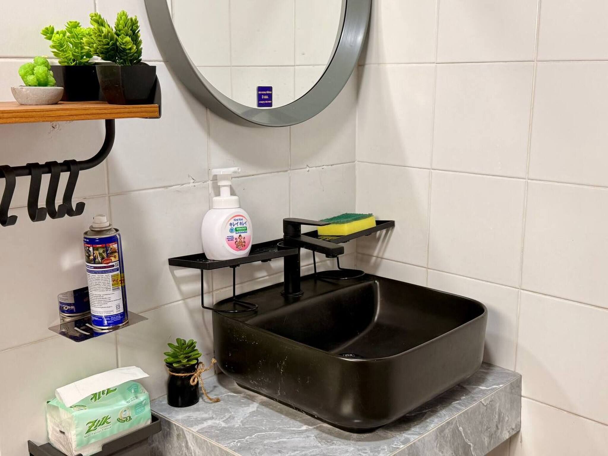 A bathroom with a black sink, a mirror, a shelf with plants, a soap dispenser, and a sponge.