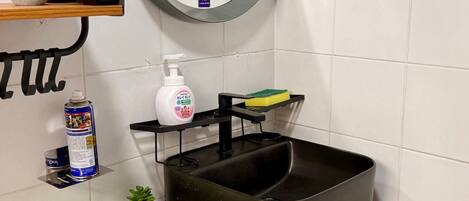 A bathroom with a black sink, a mirror, a shelf with plants, a soap dispenser, and a sponge.