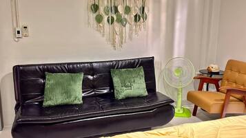 A room with a black leather sofa, two green cushions, a yellow bedspread, a green standing fan, and a small table with a decorative item.