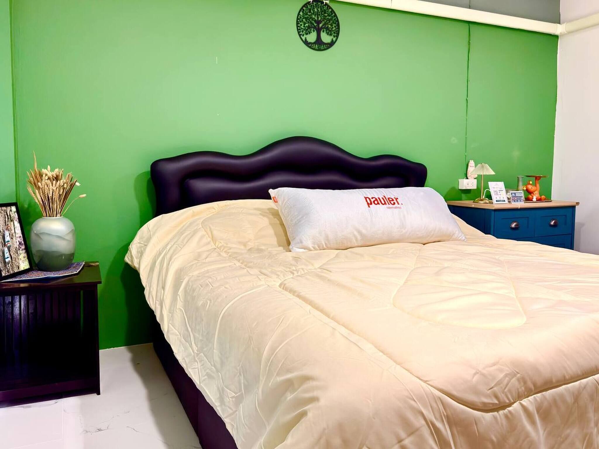 A bed with a white pillow labeled 'pauler,' a dark headboard, a green wall, a small bedside table with a lamp, and a vase with dried plants.