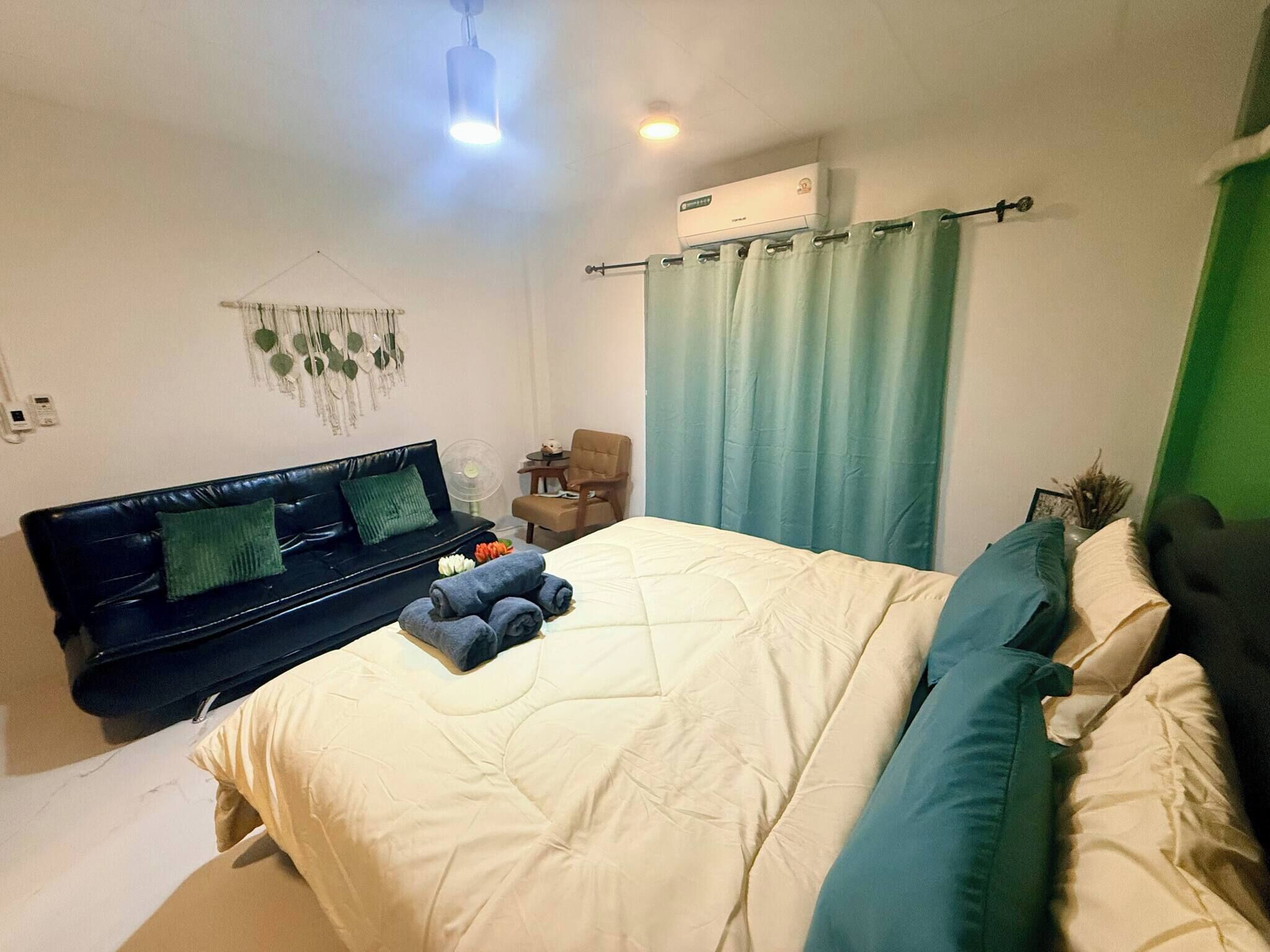 A bedroom with a bed, a black sofa, a chair, a wall-mounted air conditioner, and a ceiling fan.