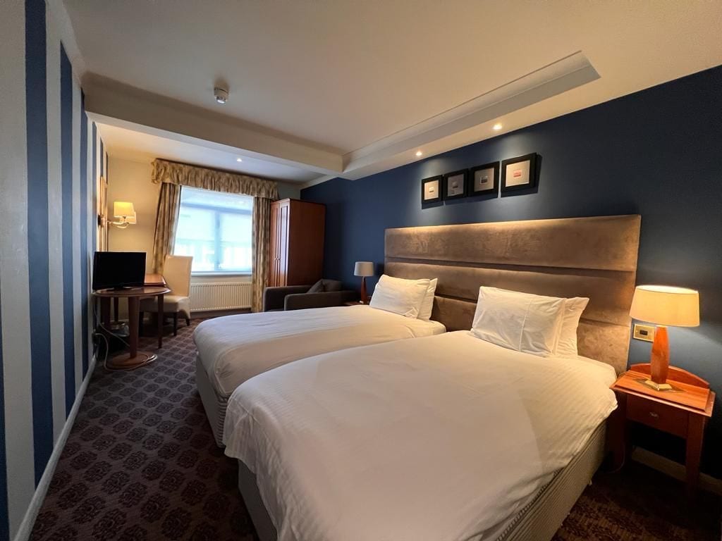 A hotel room with two beds, a desk, a chair, a television, and a window with curtains.