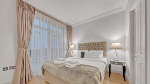 Ein Schlafzimmer mit einem großen Bett, zwei Nachttischen mit Lampen, einem Fenster mit durchsichtigen Vorhängen und einer Tür.