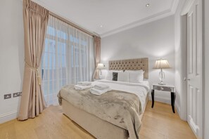 Ein Schlafzimmer mit einem großen Bett, zwei Nachttischen mit Lampen, einem Fenster mit durchsichtigen Vorhängen und einer Tür.