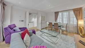 Ein modernes Wohnzimmer mit einem violetten Sofa, einem grauen Sofa und einem Glastisch. Es gibt eine Essecke mit Tisch und Stühlen, und im Hintergrund befindet sich eine Küche.