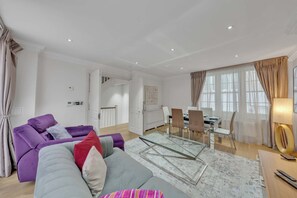 Ein modernes Wohnzimmer mit einem violetten Sofa, einem grauen Sofa und einem Glastisch. Es gibt eine Essecke mit Tisch und Stühlen, und im Hintergrund befindet sich eine Küche.