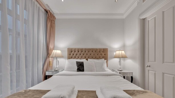 Ein Hotelzimmer mit einem großen Bett, zwei Nachttischlampen, einem gebürsteten Kopfteil und Blick auf die Stadt durch große Fenster.