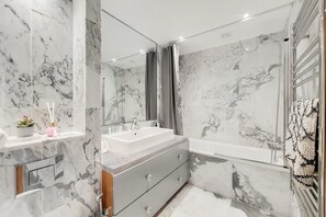 Ein modernes Badezimmer mit Marmorwänden, einem großen Spiegel, einem Waschbecken und einer Badewanne.