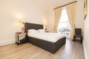Ein Schlafzimmer mit einem großen Bett, zwei Nachttischen, einem Sessel und einem Fenster mit Vorhängen.