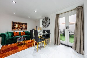 A modern living room with a green sofa, dining table, and chairs, a painting, and a view through a glass door. - Deluxe 1 Bedroom Apartment (London)