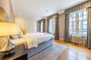 A modern bedroom with a large bed, a bedside table, a lamp, and a view of the city through the windows. - Royal Two Bedroom Apartment (London)