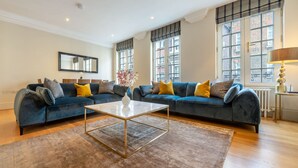 A modern living room with a blue sofa set, a white coffee table, and a large window with a view of buildings. - Royal Two Bedroom Apartment (London)