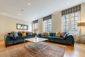 A modern living room with a blue sofa set, a white coffee table, and a large window with a view of buildings. - Royal Two Bedroom Apartment (London)