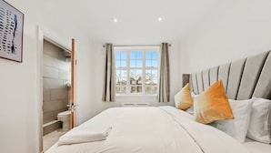 Ein Schlafzimmer mit Bett, Kissen, einem Fenster mit Vorhängen, einer Tür zu einem Badezimmer und einem gerahmten Bild an der Wand.