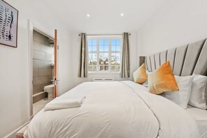 Ein Schlafzimmer mit Bett, Kissen, einem Fenster mit Vorhängen, einer Tür zu einem Badezimmer und einem gerahmten Bild an der Wand.