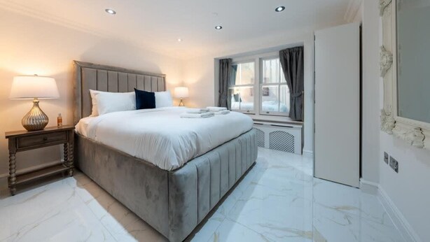 A hotel room with a large bed, bedside lamps, a window with curtains, and a radiator. - Cozy Haven Suite in London (London)