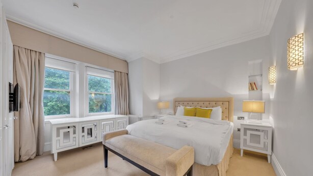 Ein Hotelzimmer mit einem großen Bett, zwei Nachttischen, einer Bank, einem Fenster mit Vorhängen und einer Wandleuchte.