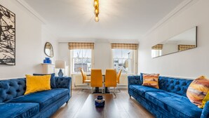 A living room with two blue sofas, a coffee table, and a dining area with yellow chairs. - Super Deluxe 2bedroom Apartment in Mayfair (London)