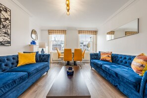 A living room with two blue sofas, a coffee table, and a dining area with yellow chairs. - Super Deluxe 2bedroom Apartment in Mayfair (London)