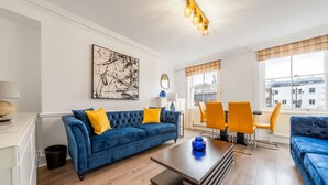 A modern living room with a blue sofa, yellow cushions, a coffee table, and a dining area with orange chairs. - Super Deluxe 2bedroom Apartment in Mayfair (London)