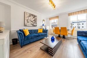 A modern living room with a blue sofa, yellow cushions, a coffee table, and a dining area with orange chairs. - Super Deluxe 2bedroom Apartment in Mayfair (London)