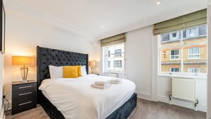 A hotel room with a large bed, a nightstand, a lamp, and a window with a view of a building. - Super Deluxe 2bedroom Apartment in Mayfair (London)