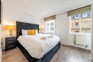 A hotel room with a large bed, a nightstand, a lamp, and a window with a view of a building. - Super Deluxe 2bedroom Apartment in Mayfair (London)