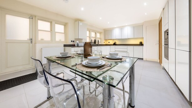 A modern kitchen with a glass dining table, clear chairs, and white cabinetry. - Exquisite 3-bedroom House in Mayfair (London)