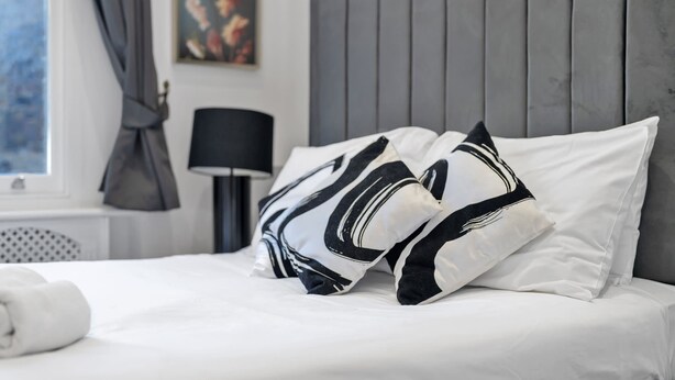 Ein Bett mit weißer Bettwäsche und abstrakten Kissen in Schwarz-Weiß, eine schwarze Lampe und ein grauer Vorhang.
