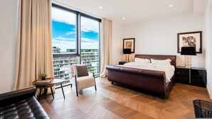 A modern hotel room with a large bed, a seating area with a chair and a small table, a side table with a lamp, and a view of the cityscape through large windows. - Luxury 4-bed Duplex Penthouse in Battersea (London)