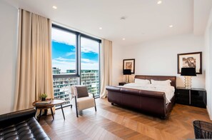 Ein modernes Hotelzimmer mit einem großen Bett, einer Sitzecke mit einem Sessel und einem kleinen Tisch, einem Nachttisch mit Lampe sowie Blick auf die Stadt durch große Fenster.