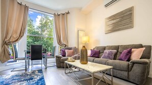 A living room with a sofa, a coffee table, a lamp, a chair, and a view through a large window. - Exclusive 3-bedroom Apartment (London)