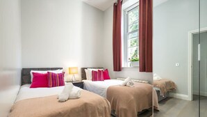A hotel room with two beds, a window with curtains, a bedside table with a lamp, and a door. - Exclusive 3-bedroom Apartment (London)