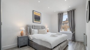 A modern bedroom with a large bed, two bedside tables, a window with curtains, and a painting on the wall. - Standard 1 Bedroom Apartment In Belgravia (London)
