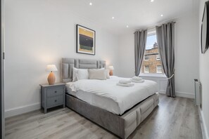 A modern bedroom with a large bed, two bedside tables, a window with curtains, and a painting on the wall. - Standard 1 Bedroom Apartment In Belgravia (London)