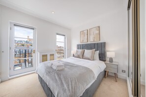 A bedroom with a large bed, a nightstand, a window with a view of buildings, and two wall-mounted artworks. - Modern 4-bed Apartment in Knightsbridge (London)