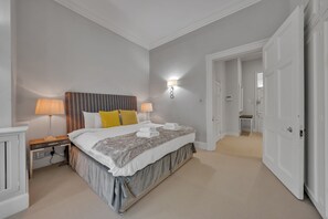 Ein Schlafzimmer mit einem Bett, Nachttischlampen, einem Beistelltisch und einer Tür zu einem anderen Raum.