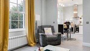 A modern living room with a grey armchair, a window with yellow curtains, and a view of the outdoors. - Superior 3 Bedroom Apartment (London)