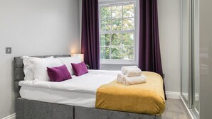 A neatly made bed with white linens and two purple pillows, a mustard-colored throw blanket, and a window with purple curtains. - Superior 3 Bedroom Apartment (London)